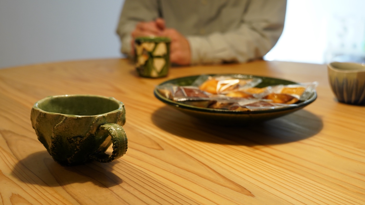 木のテーブルに並ぶ緑のうつわとお菓子、カップに手を添える人